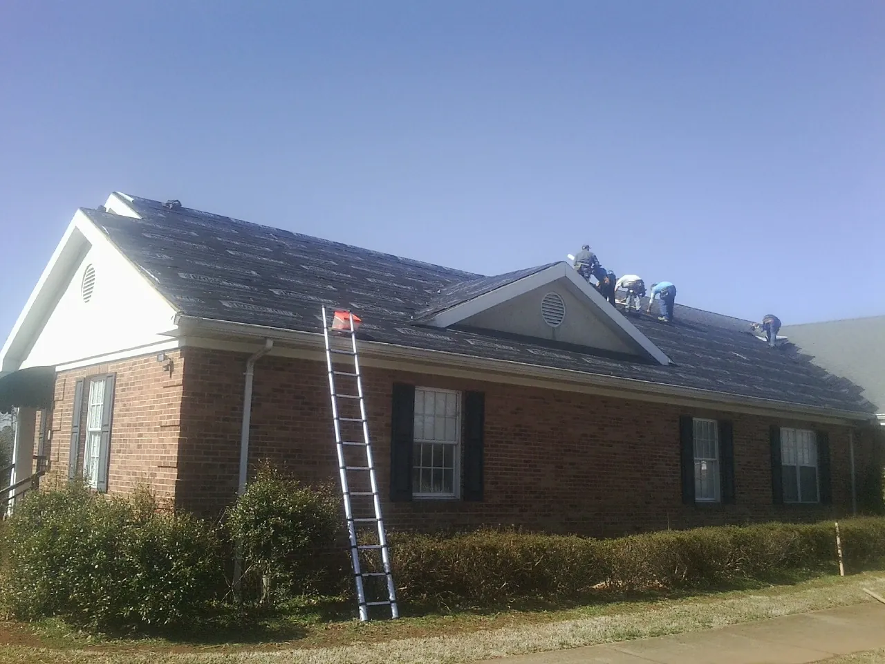 Crew tearing shingles off a brick ranch roof with a ladder set — Emergency Roof Repair work by our crew in Neenah