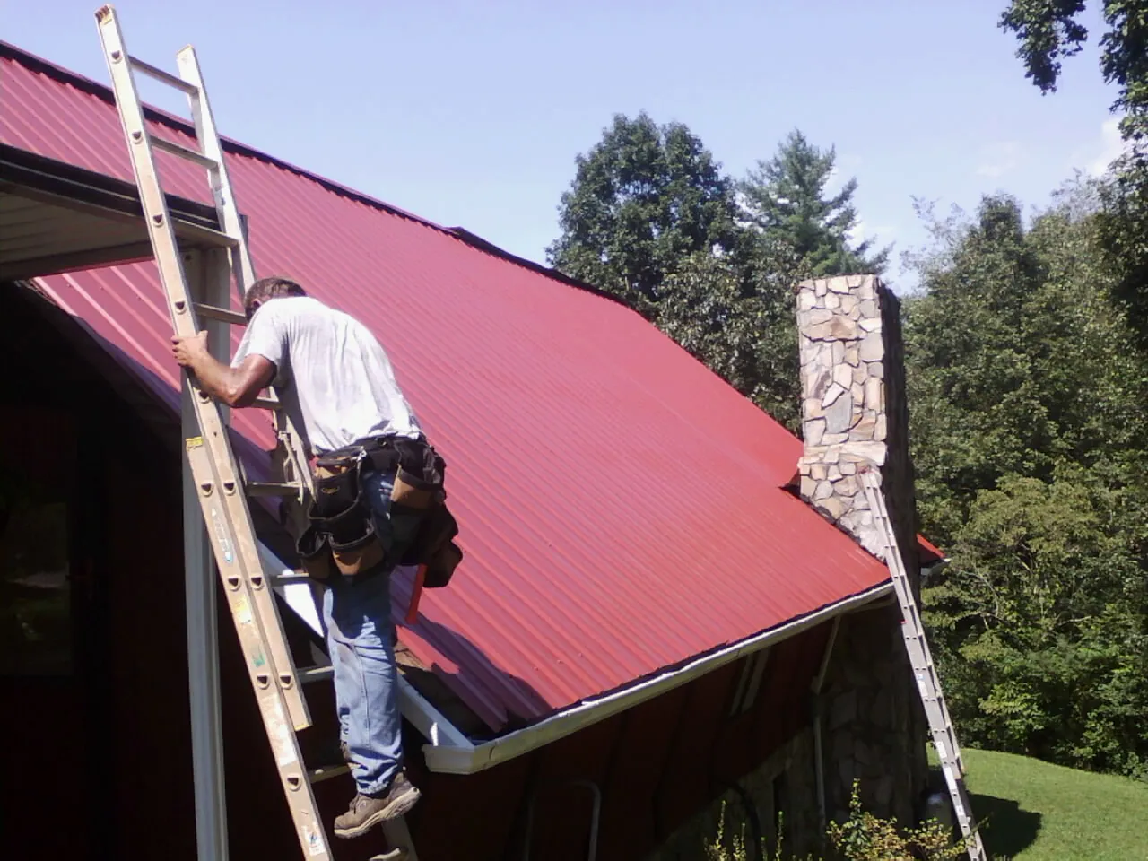 Roofer on a ladder beside a red metal roof and stone chimney — Soffit & Fascia Repair by Roofing Brothers in Neenah